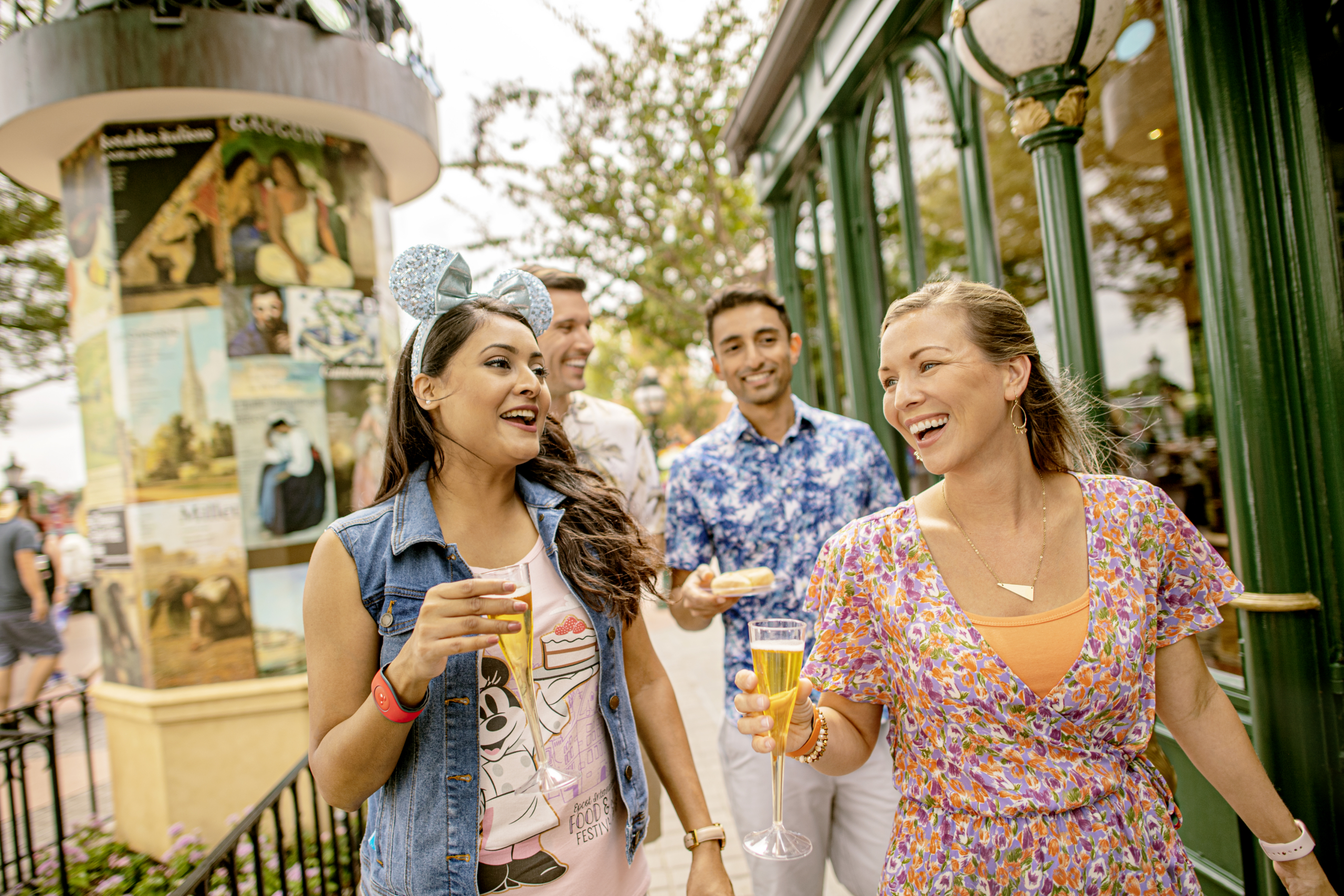 Four friends smile and chat while holding drinks at an outdoor event near a green fence and colorful mural.