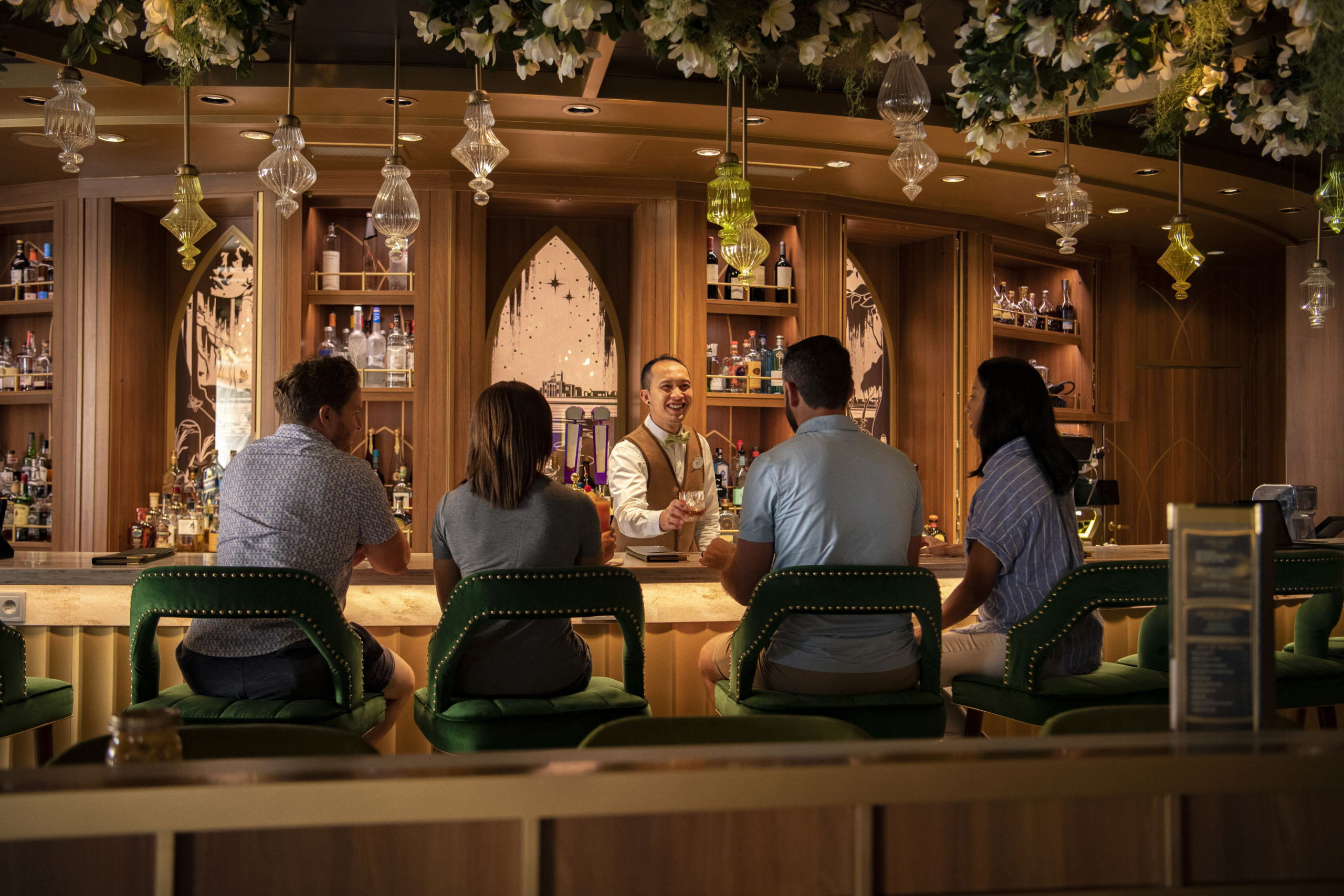 A bartender in a vest serves drinks to four patrons seated at a stylish bar with wooden shelves and hanging glass