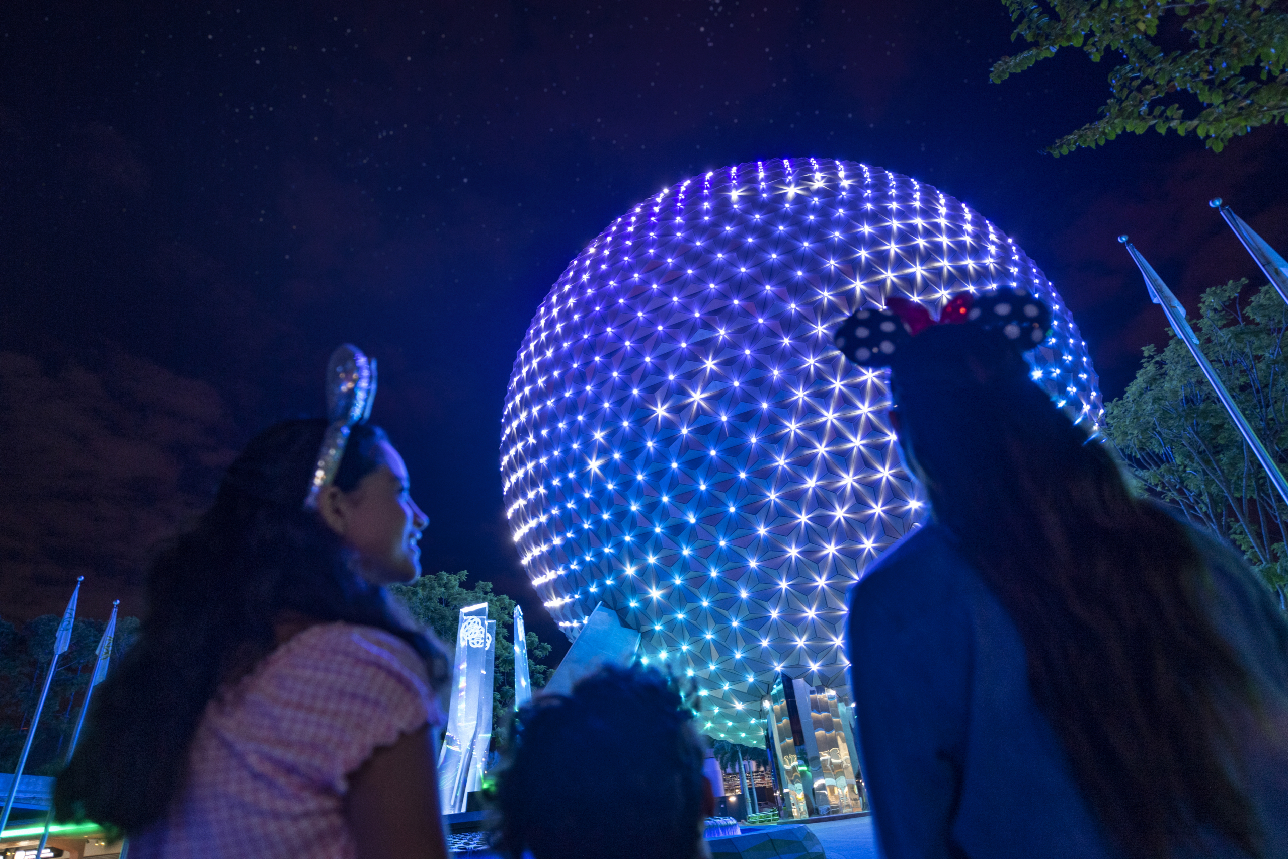 Two women and a child look up at the illuminated Geodesic Sphere at night, surrounded by trees and flags.