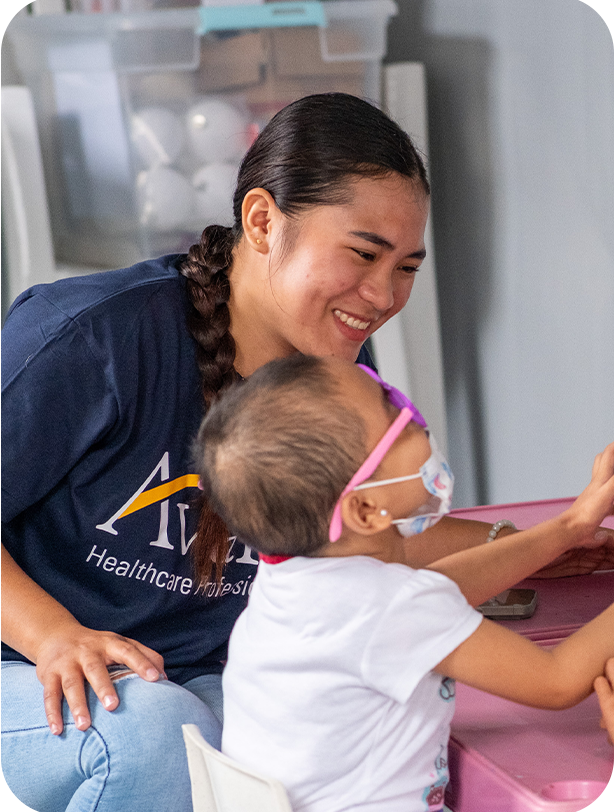 A healthcare professional smiles as she interacts with a young girl wearing a face mask and glasses, sitting at a pink table.