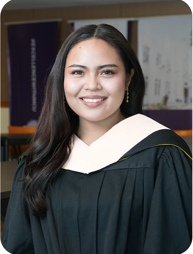 A young woman in a graduation gown and white hood smiles indoors, with a blurred background of banners and posters.