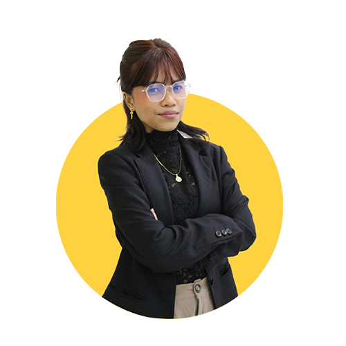 A woman with glasses, earrings, and layered jewelry, wearing a black blazer and standing confidently with arms crossed