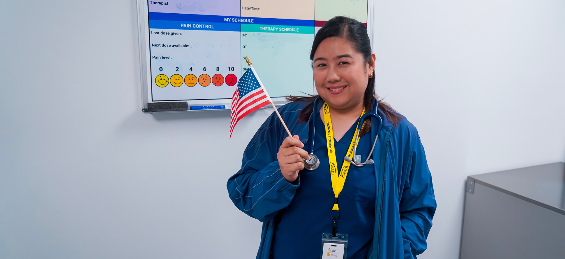 A smiling healthcare professional holding an American flag, wearing a blue uniform and stethoscope, standing in front of a