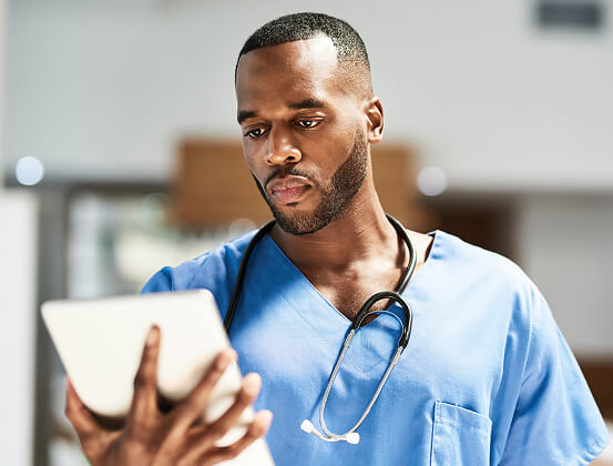 A healthcare professional in blue scrubs with a stethoscope looks at a tablet device.