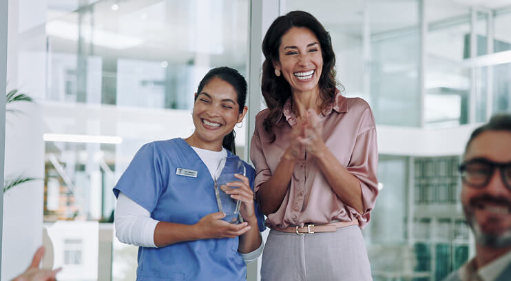 Two healthcare professionals, one in scrubs holding an award and another smiling, celebrate in a modern hospital setting.