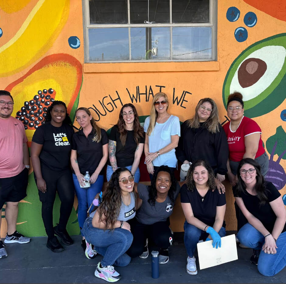 A diverse group of people stands in front of a colorful mural with fruit illustrations, smiling and celebrating together.