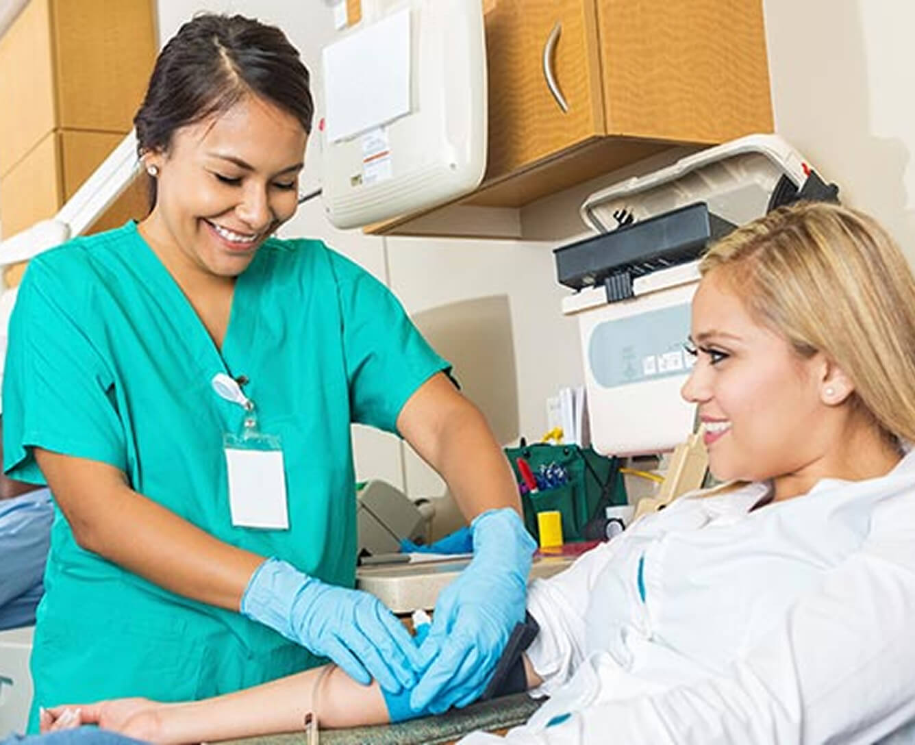 A nurse in teal scrubs draws blood from a smiling patient lying in a hospital bed.