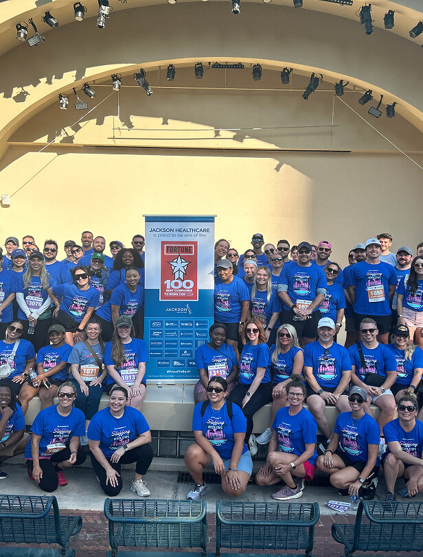 A large group of people in matching blue shirts pose together at a charity event, with a Jackson Healthcare banner behind