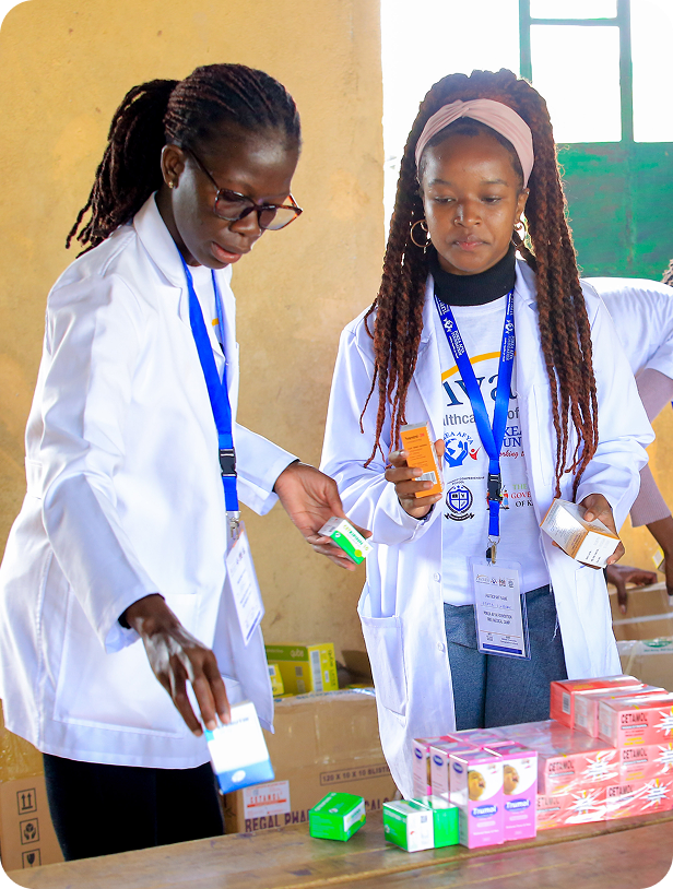 Two women in white lab coats examine and compare medication boxes at a table during a health-related event.