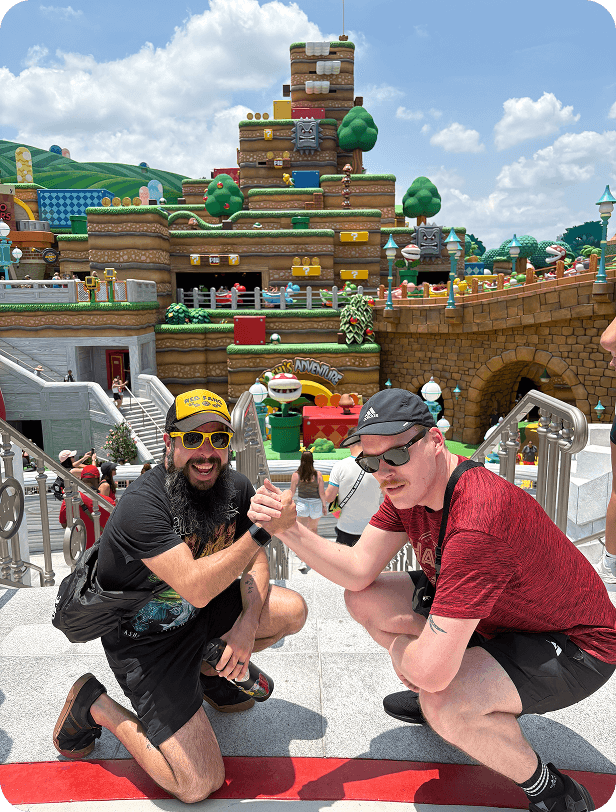 Two men arm-wrestle in front of a colorful Super Mario-themed amusement park with a large castle structure in the background.