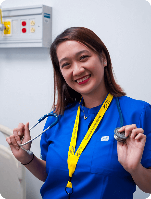 A smiling nurse in blue scrubs holds a stethoscope in one hand and a penlight in the other, wearing a yellow lanyard with