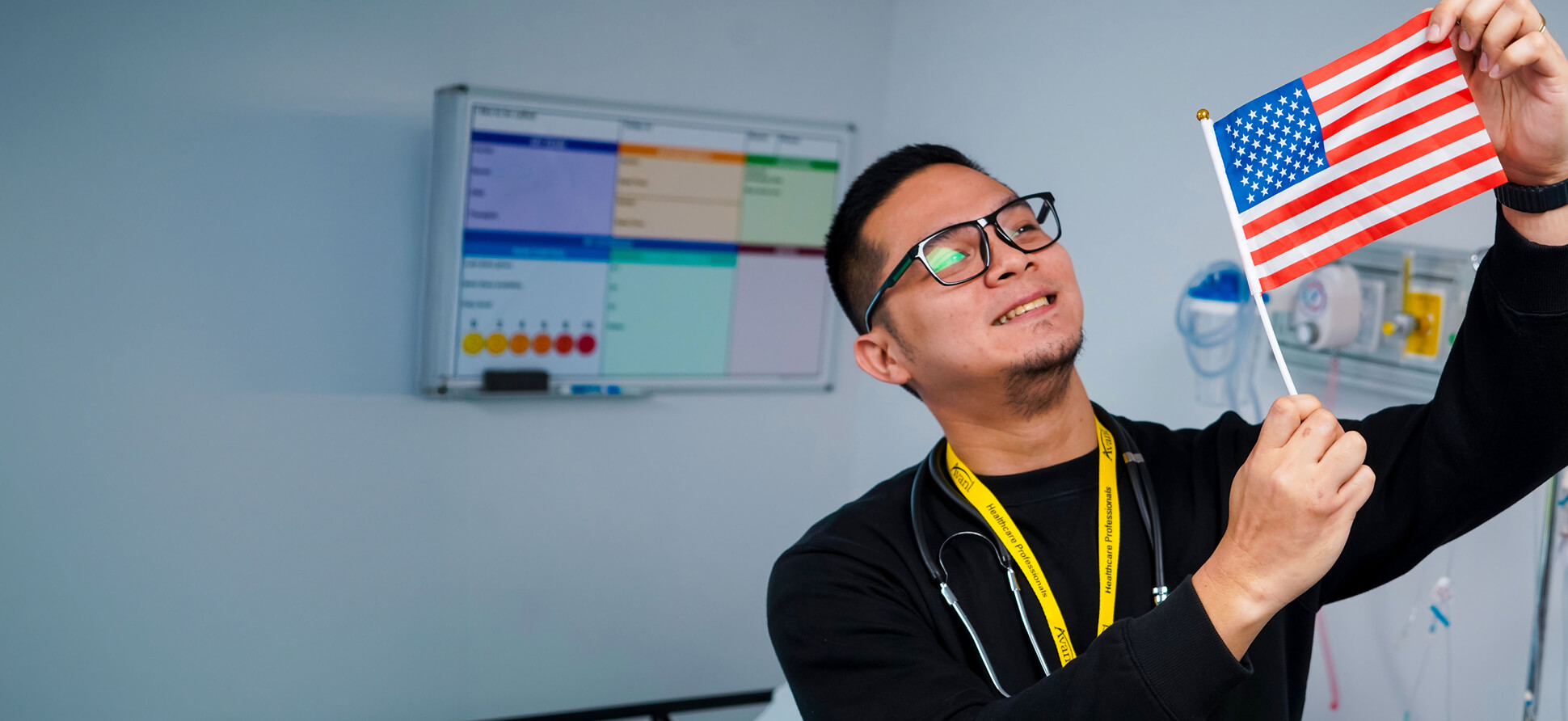 A healthcare professional holding an American flag in a hospital room with a colorful medical chart on the wall.