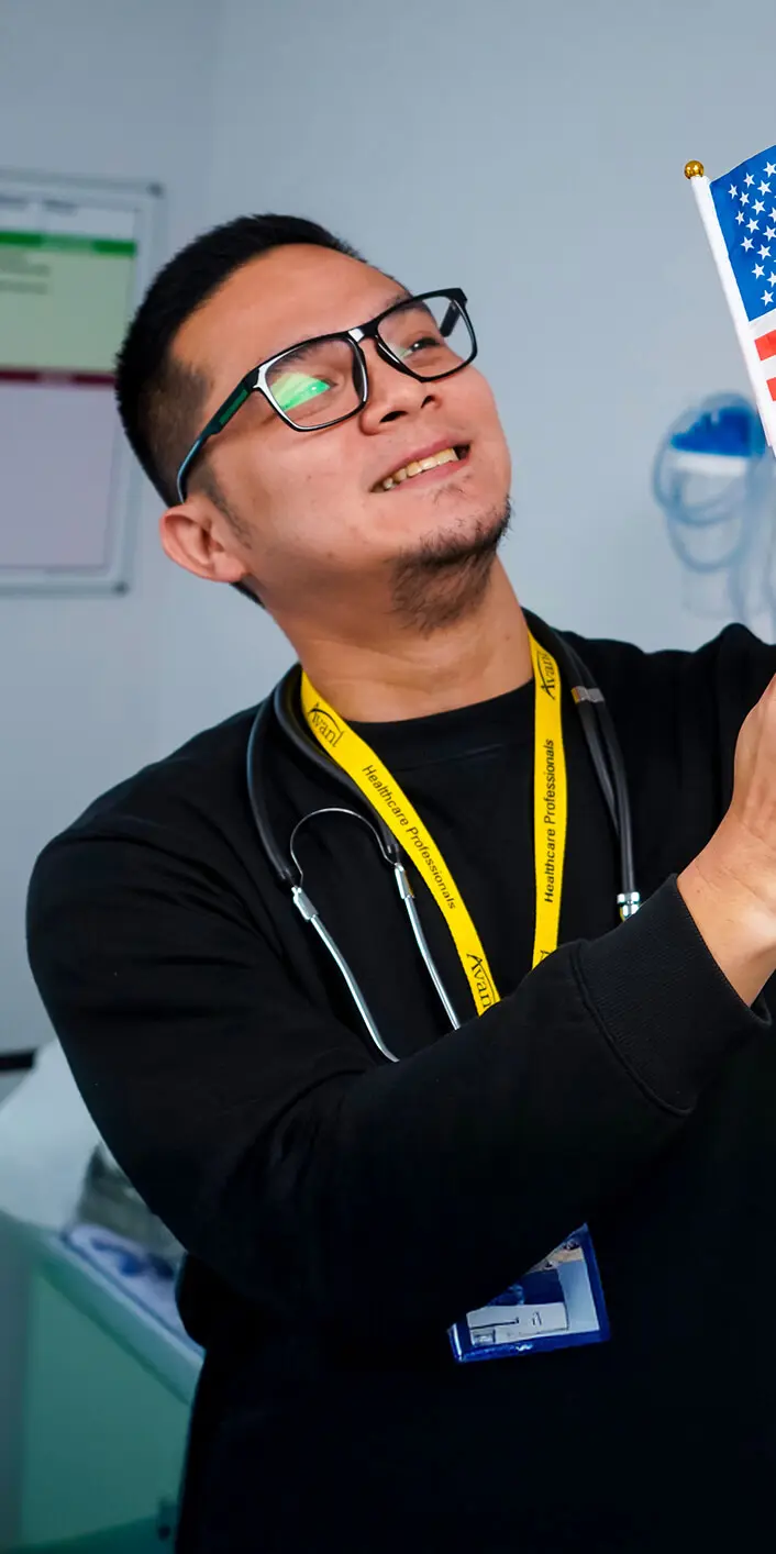 A smiling healthcare professional wearing glasses, a stethoscope, and a yellow lanyard, holding a small American flag.