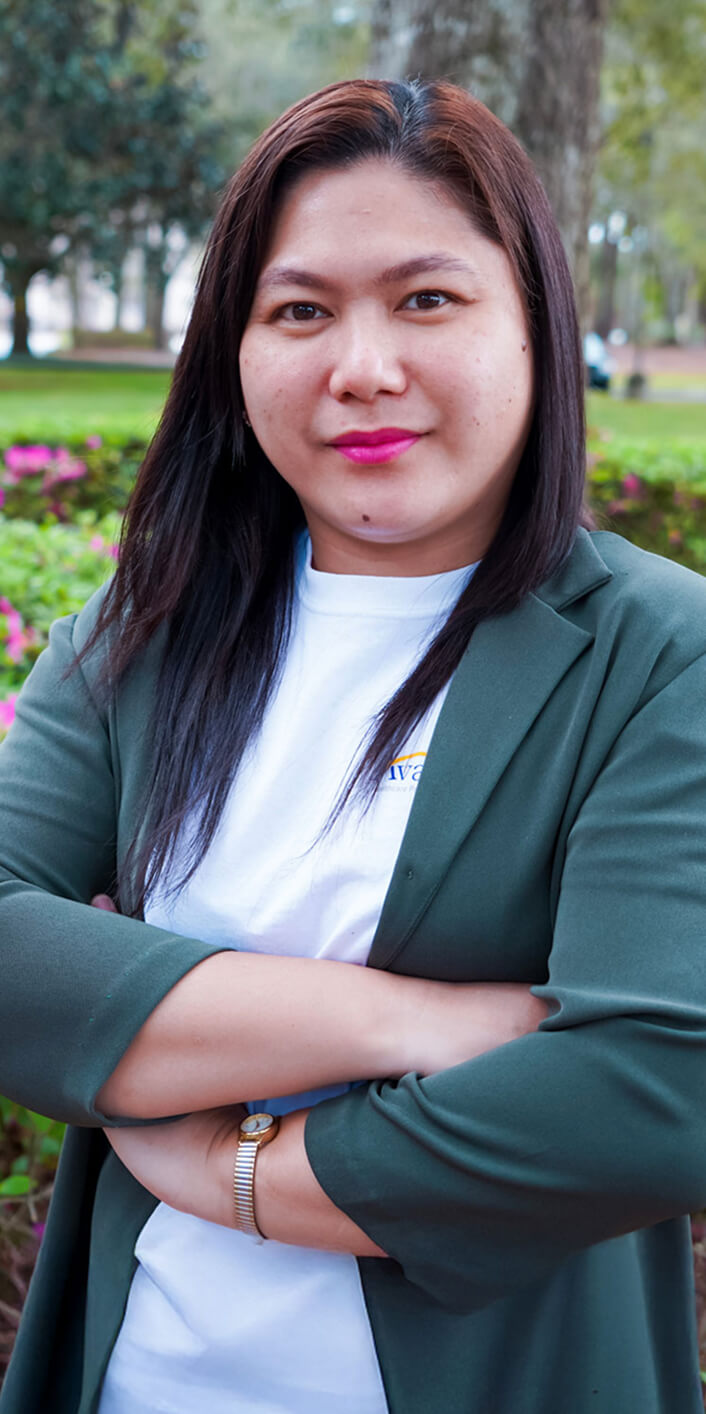 A woman with long dark hair, wearing a green blazer and white shirt, stands outdoors with arms crossed in a park.
