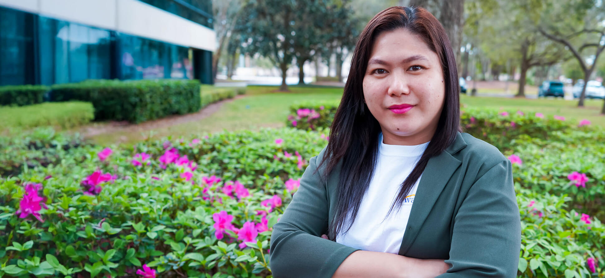 A woman with long dark hair and pink lipstick stands outdoors among pink flowers and green bushes, with trees and a building