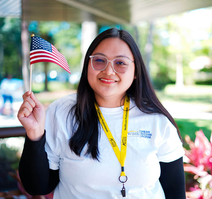 A smiling woman with glasses holds an American flag, wearing a white T-shirt and a yellow lanyard, outdoors with greenery in
