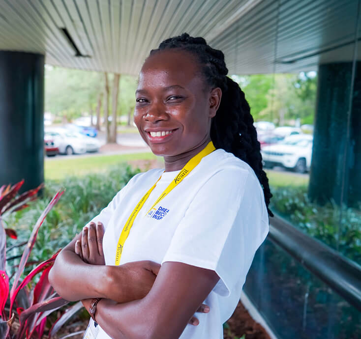 A smiling woman with braided hair wearing a white shirt and yellow lanyard stands outdoors with arms crossed, promoting good
