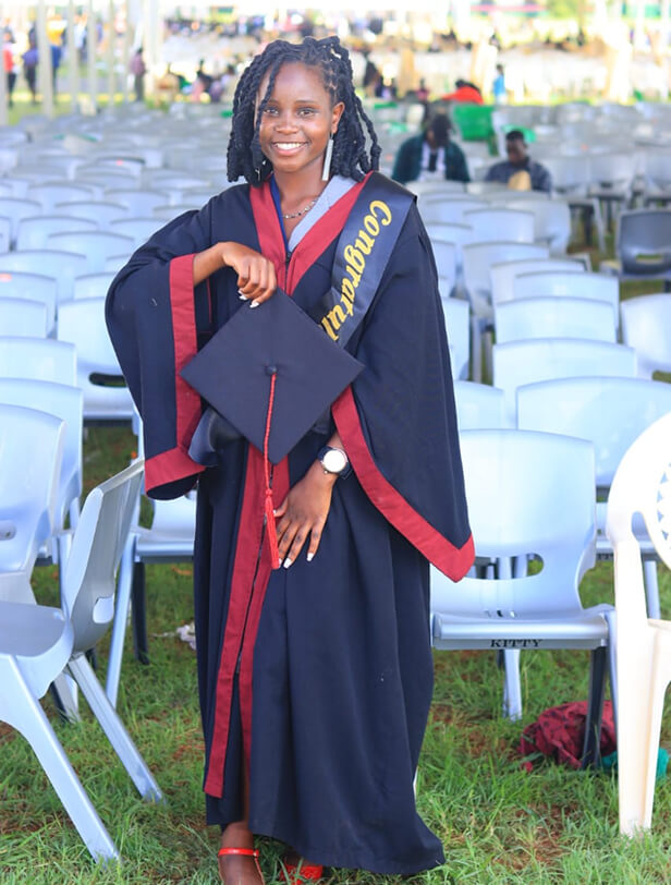 A young woman in a graduation gown and cap stands outdoors among empty chairs, smiling with her diploma in hand.