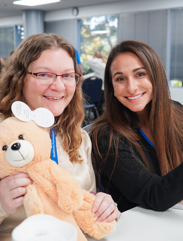Two women smiling at a conference, one holding a plush teddy bear, with a bright, modern office background.