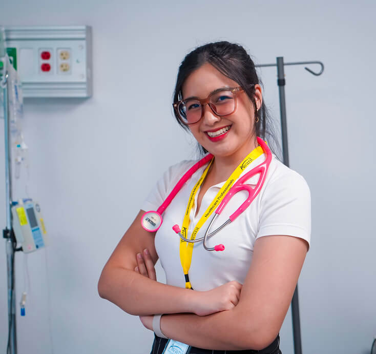 A smiling female healthcare professional with glasses and a pink stethoscope around her neck stands confidently in a medical