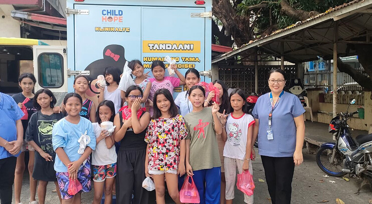 Group of children and a woman in medical scrubs posing outdoors in front of a mobile health unit.
