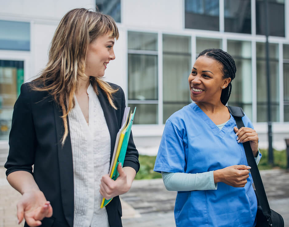 A nurse in blue scrubs smiling and talking with a woman holding folders outside a modern building.