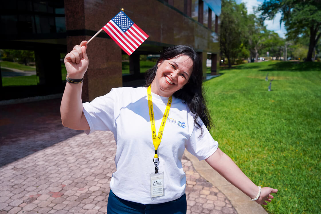 A smiling woman with dark hair holding an American flag outdoors, wearing a white T-shirt and a yellow lanyard.