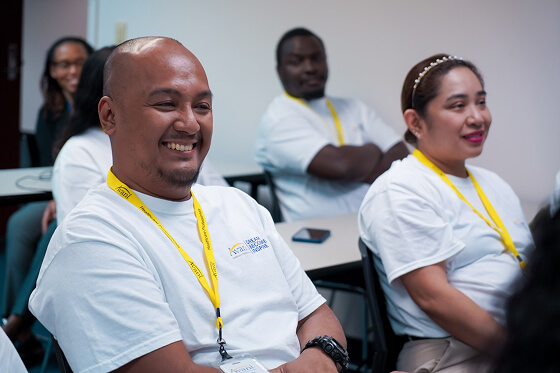 Participants wearing white shirts and yellow lanyards sit and smile during a training session.
