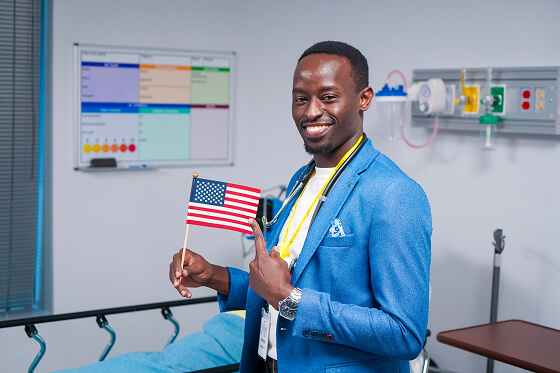 A smiling man in a blue suit holds an American flag and peace sign in a hospital room with medical equipment and a schedule