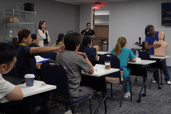 A diverse group of candidates attends a CPR training class, watching an instructor demonstrate on a mannequin.