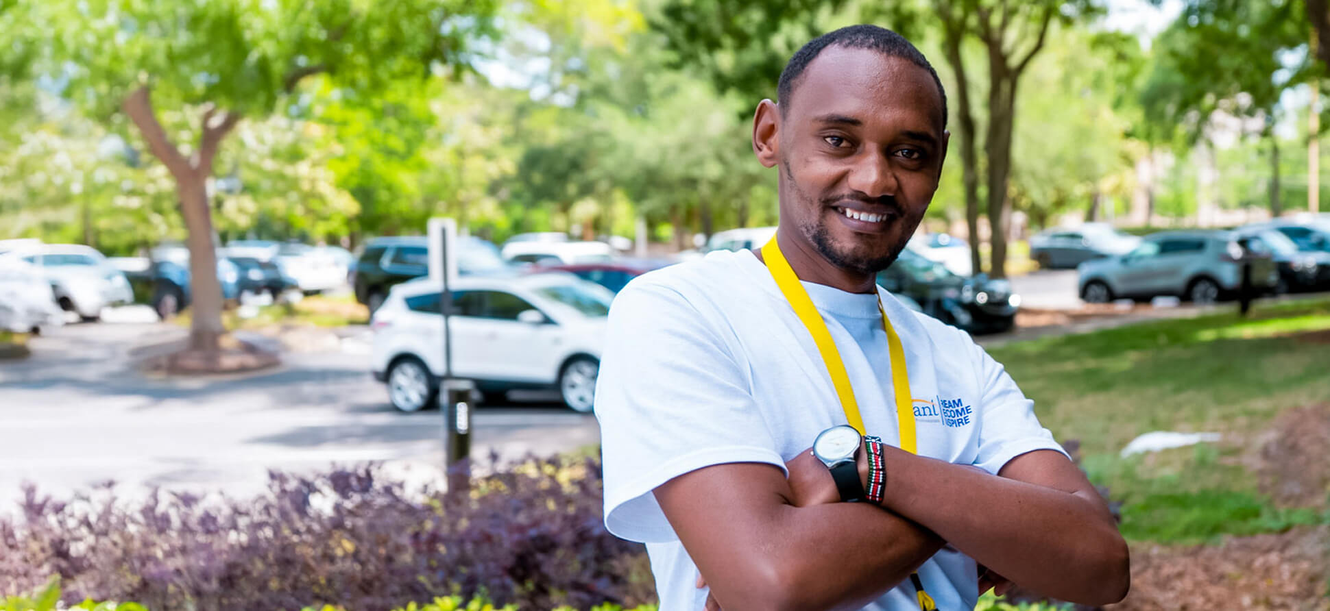 A smiling man with crossed arms wearing a white T-shirt and a yellow lanyard outdoors in a parking lot with green trees.