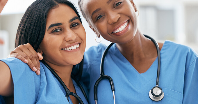 Two smiling healthcare professionals in blue scrubs with stethoscopes around their necks share a friendly moment.