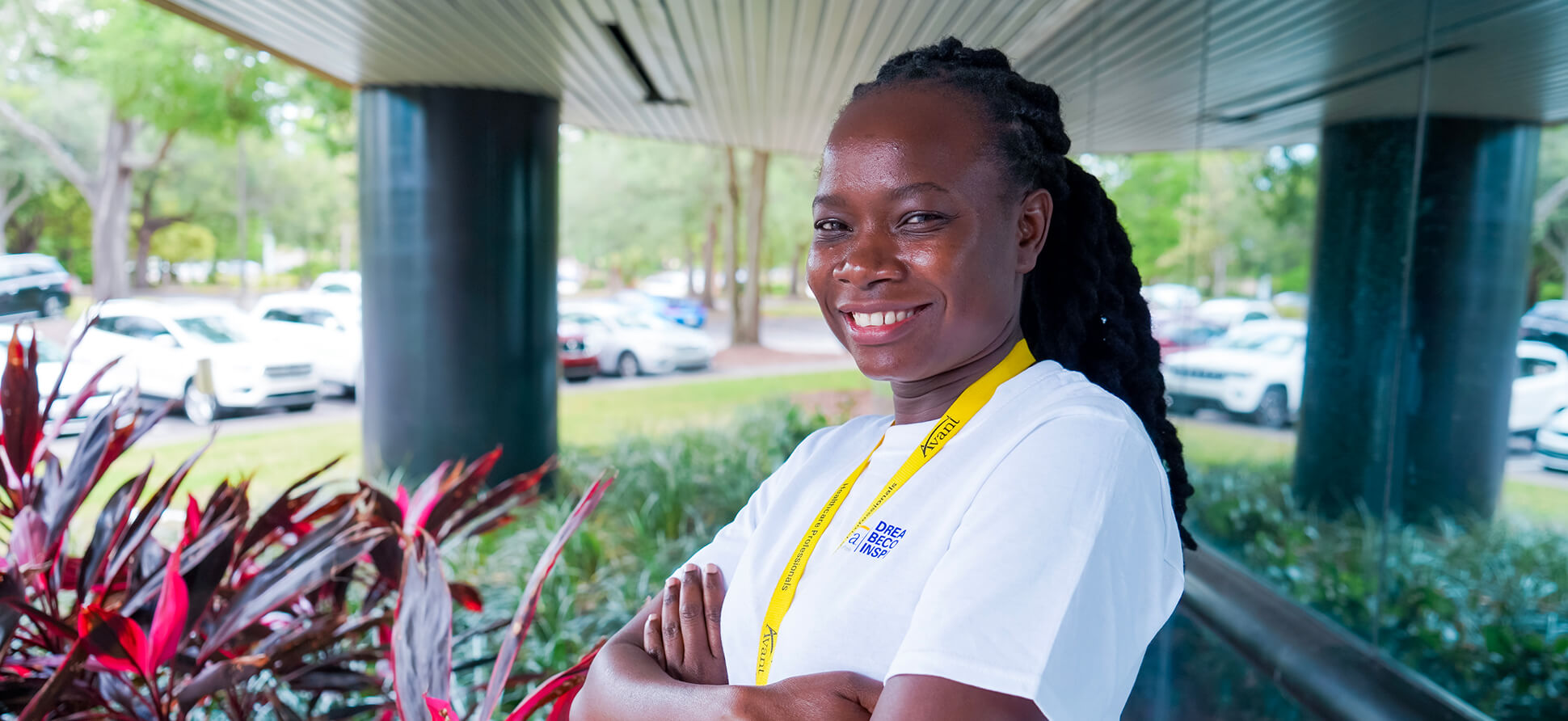 A smiling woman with braided hair wearing a white shirt and a yellow lanyard stands outdoors near red plants and parked cars.
