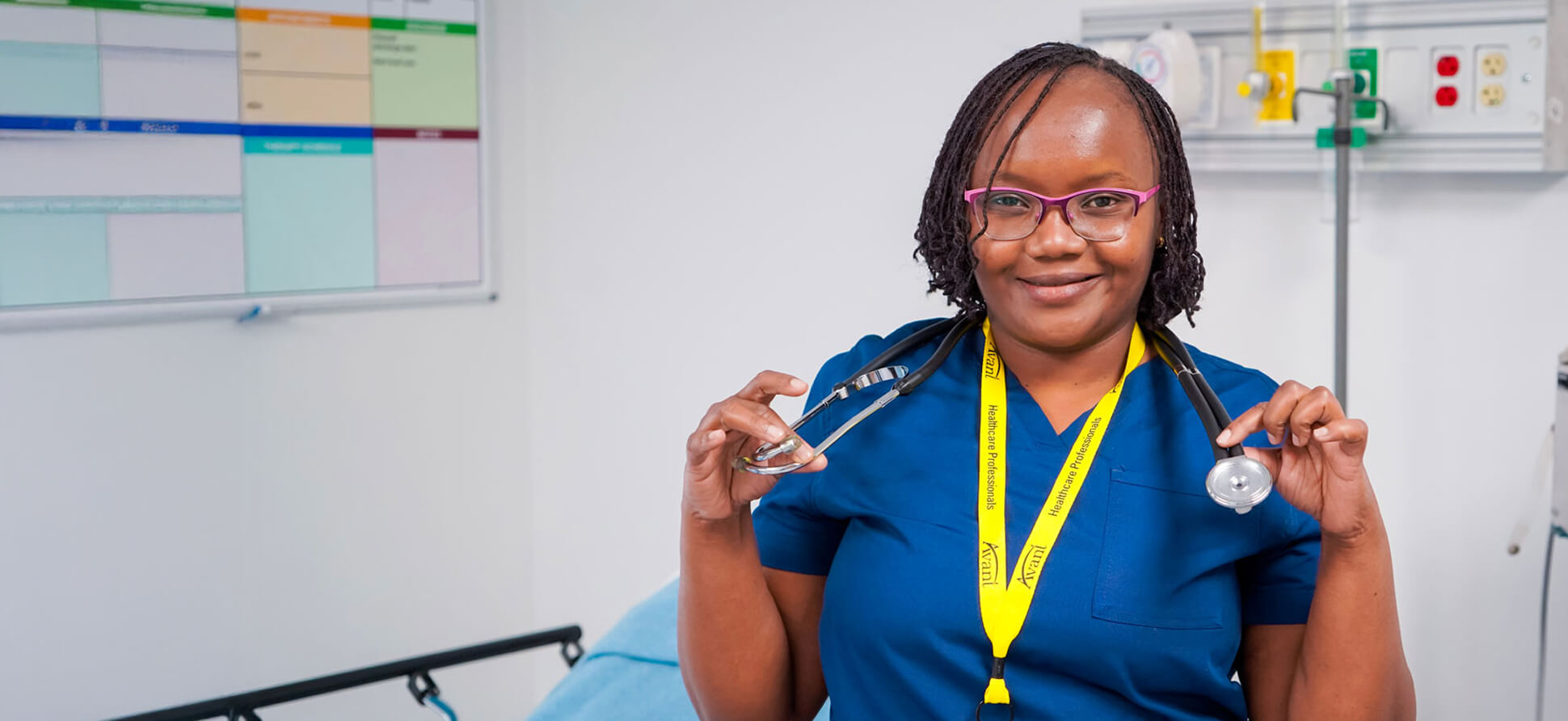 A smiling healthcare professional in blue scrubs holding a stethoscope in a medical facility in Kenya.