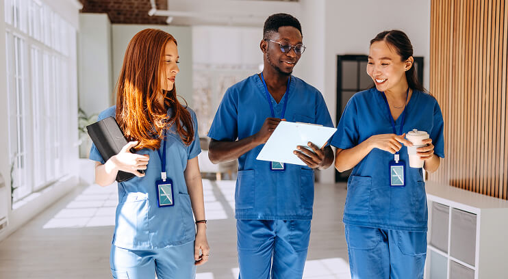 Three diverse nurses in blue scrubs walk and chat in a bright hospital corridor, smiling and holding medical supplies.