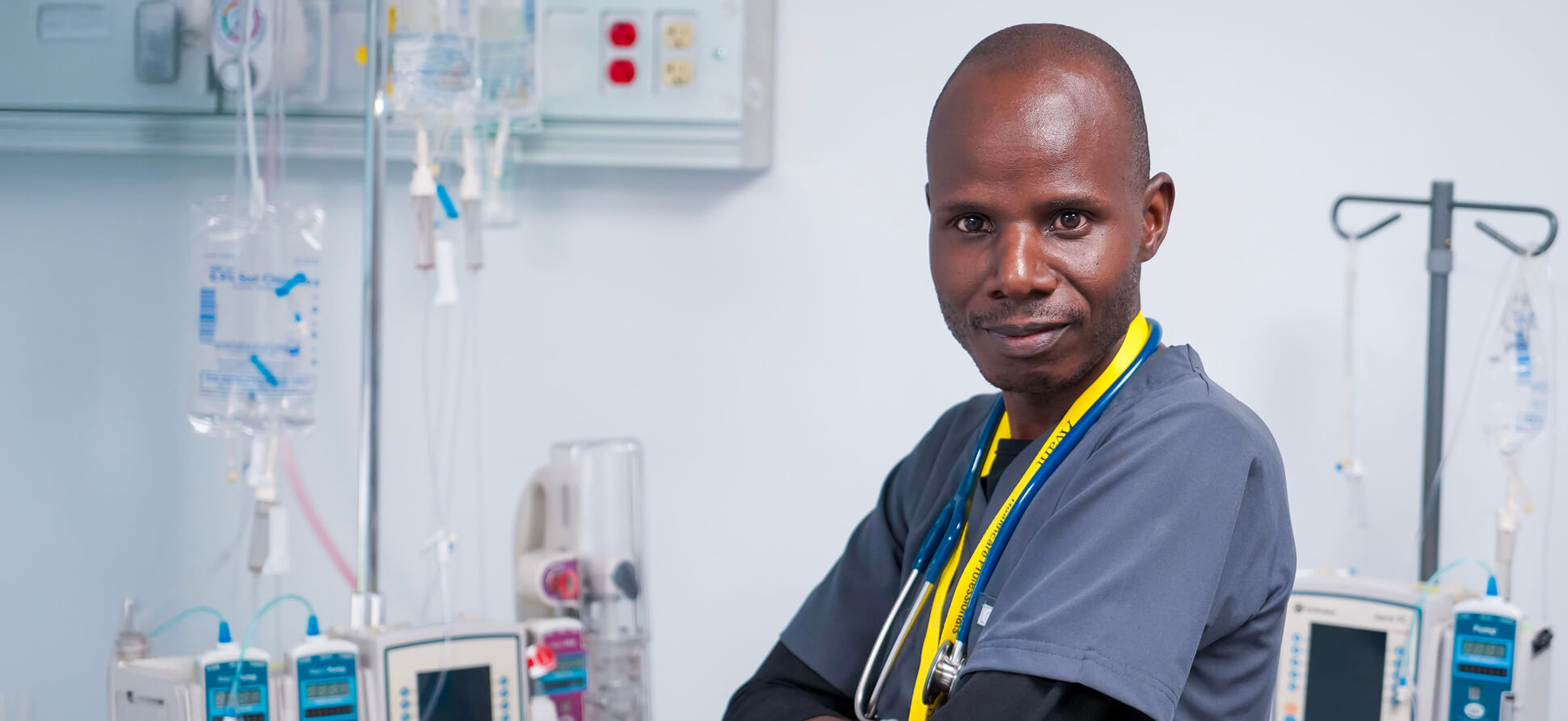 A Kenyan healthcare professional in scrubs with a stethoscope around his neck, standing in a hospital ICU with medical