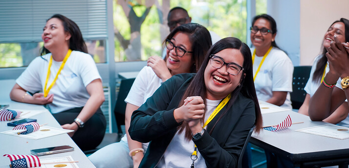 Avant-Our-International-Nurse-Program-Image@2x-1 Group of diverse smiling nurses in a classroom, wearing white uniforms and yellow lanyards, engaged in a training session.