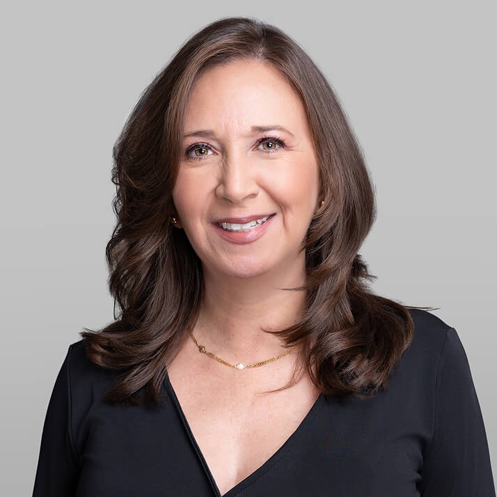 Ada DeGuzman with shoulder-length brown hair, wearing a black top and a delicate gold necklace, smiling against a gray