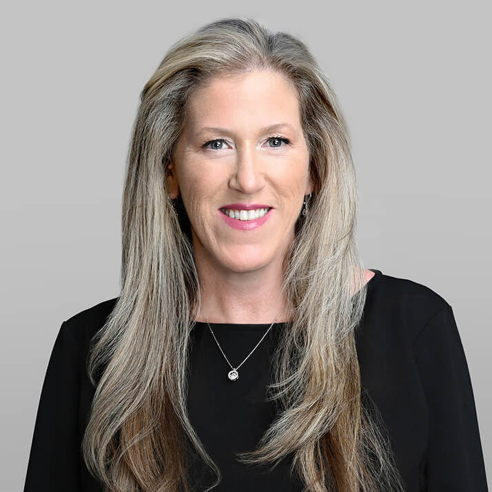 Brenda Kuczynski with long blonde hair, wearing a black top and a silver necklace, smiling against a gray background.