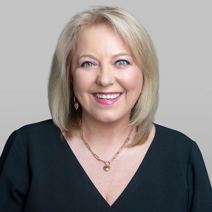 Lesley Hamilton-Powers with blonde hair, wearing a black top and gold jewelry, smiling against a light gray background.