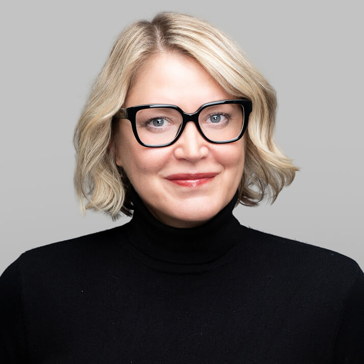 Marisa Zaharoff with blonde hair, black glasses, and a black turtleneck, smiling against a gray background.