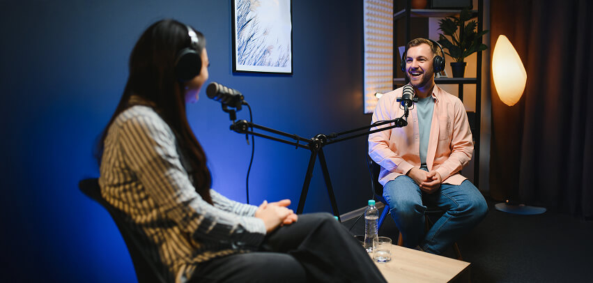 Avant Podcast Hero A man and woman wearing headphones sit in a recording studio, smiling and speaking into microphones during a podcast episode.