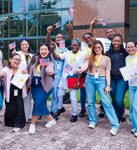 Avant Program Overview Hero1 A diverse group of smiling people celebrating outdoors with American flags and certificates in front of a modern building.
