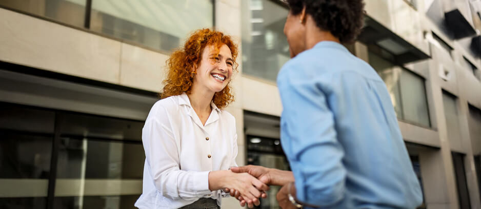 A woman with curly red hair shakes hands with a person in a blue shirt outside a modern building, smiling warmly.