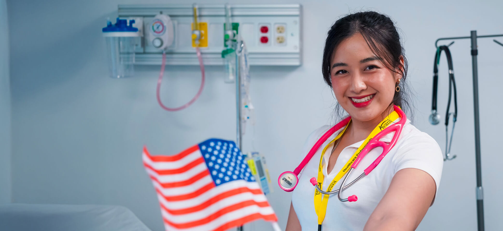 A smiling female healthcare worker with a pink stethoscope around her neck holds an American flag in a hospital room.