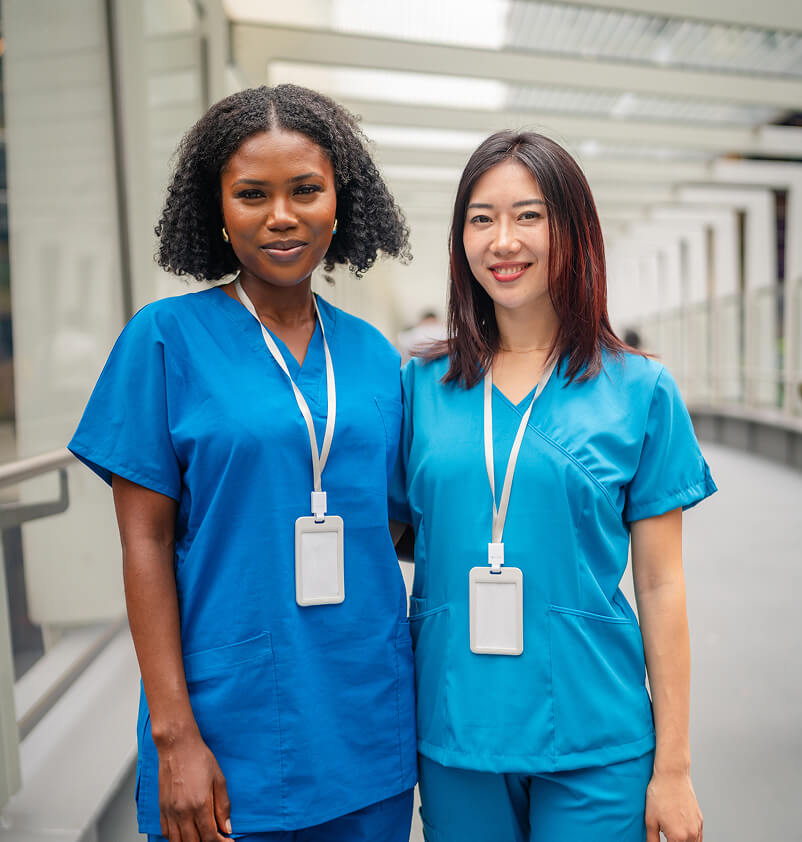 Two diverse healthcare professionals in blue scrubs stand together in a modern hospital corridor, smiling confidently.