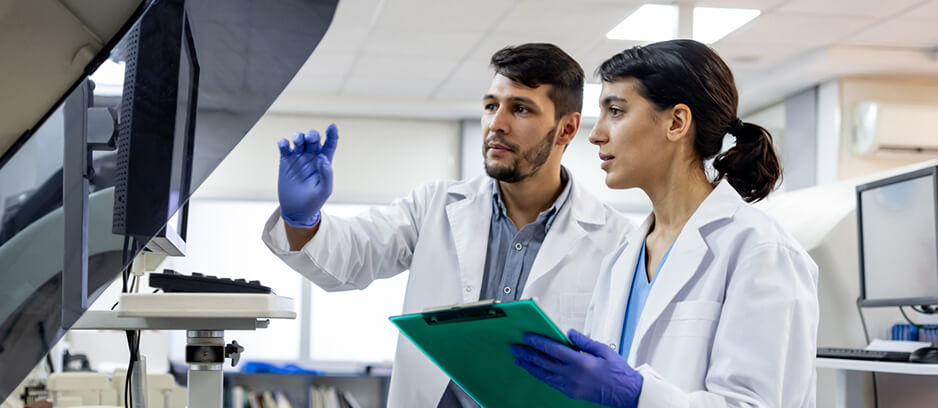 Two medical technologists in lab coats and gloves review data on a computer screen and clipboard in a clinical laboratory.