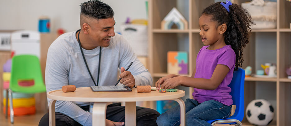 A therapist and young girl sit at a table engaging in a therapy session with toys and a tablet in a colorful playroom.