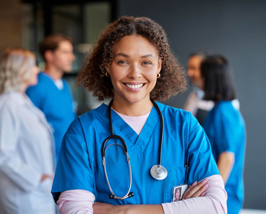 A smiling female healthcare professional in blue scrubs with a stethoscope, standing confidently with colleagues in the
