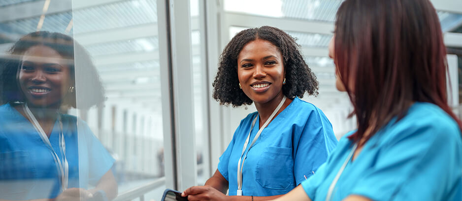 Two smiling registered nurses in blue scrubs conversing in a bright hospital corridor.